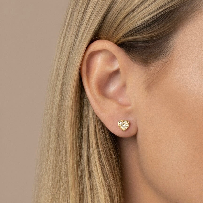 Gold heart-shaped earring worn by a person with blonde hair against a beige background